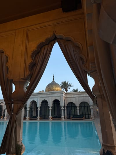 The image shows a view through an ornate doorway of a building, with a swimming pool and a dome-shaped structure in the background. The building is made of white stone with intricately carved arches. The pool is filled with clear blue water and reflects the sky above. The dome is gold and rests on a white structure, which also has arched doorways. A palm tree stands beside the structure. The doorway itself is made of wood and has a series of arches. The view is framed by curtains that hang on either side of the doorway. The scene is located in Ouled Jelal, Morocco, at coordinates (latitude: 31.663, longitude: -7.872).