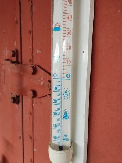 A vertical thermometer is mounted on a reddish-brown wooden surface, possibly a door or wall, in Вароніцкі сельскі Савет, Belarus. The thermometer displays temperature readings in Celsius, with markings from 0 to 50 degrees. The liquid level in the thermometer is just above the 0-degree mark, indicating a temperature slightly above freezing. Decorative winter-themed icons, such as a snowman and a Christmas tree, are visible on the thermometer. The wooden surface shows signs of age and use, with a visible hinge mechanism on the left side of the frame. The lighting suggests an indoor or shaded outdoor environment.