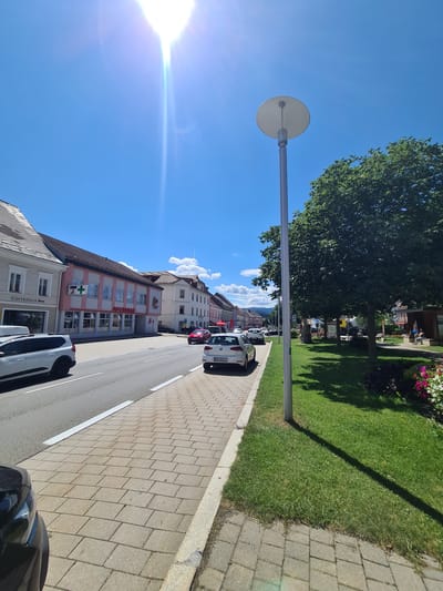 The image depicts a street scene in Sankt Marein bei Neumarkt, Austria, on a bright, sunny day with a clear blue sky and a few white clouds. The perspective is from a low angle, looking down a street lined with buildings on the left and a grassy area with trees and a lamppost on the right.

On the left side of the street, there are several buildings. The closest building on the left has a sign that reads "GÄSTEHAUS" (Guesthouse). Next to it is a building with a pink and white facade, featuring a green cross symbol and "APOTHEKE" (Pharmacy) written on it. Further down the street, more buildings are visible, with varied architectural styles and colors. Cars are parked along the street and some are driving. A white SUV is parked on the left, and several other cars, including a white Volkswagen Golf, are on the road and parked on the right side of the street.

The right side of the scene features a well-maintained green lawn with mature trees casting shadows. A modern lamppost stands prominently in the grassy area. In the background of the grassy area, there are some wooden structures, possibly benches or shelters, and some people are visible sitting or walking. A variety of flowers, including pink and red ones, are planted near the edge of the grassy area. The street itself has a paved sidewalk with interlocking stones, and a road with clear lane markings. The overall impression is of a clean and orderly urban environment.