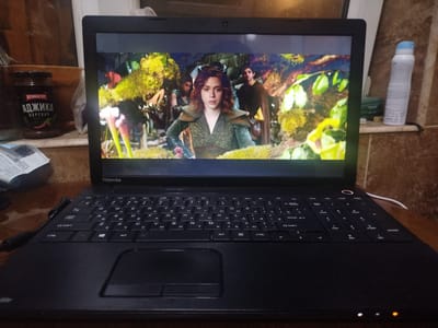 A black Toshiba laptop is open on a wooden surface. The laptop screen displays a scene from a movie or show, featuring a young woman with auburn hair wearing a green tunic, standing in a lush, natural environment with vibrant flora. In the background, two individuals are partially visible. To the left of the laptop, a jar of adjika sauce, a popular Georgian condiment, is present. Further to the left, a white piece of paper with some text is visible. To the right of the laptop, a white spray can with a blue cap stands on a shelf. The lighting suggests an indoor setting, likely a home or a room. The overall impression is a casual, personal space where someone is engaged with media content. The location is not specified in Sabunchu, Azerbaijan.