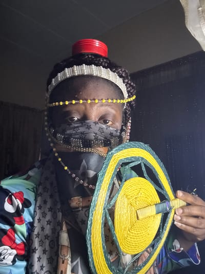A woman is holding a yellow and green woven fan. She is wearing a pearl headband, yellow beaded necklace, and a gray face covering. Her clothing includes a gray patterned shirt, and a lower layer of blue fabric with Disney characters. The location is likely a room in Madangala, Nigeria.