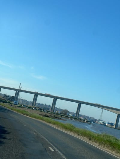 The image depicts a road running alongside a body of water, possibly a river or an estuary, under a bridge structure. The scene suggests a rural or semi-rural setting. Across the water is a settlement with buildings, trees, and what appears to be industrial or port infrastructure, including transmission towers. The sky is clear blue. The location is consistent with a landscape in Babergh, United Kingdom, characterized by waterways and bridges near urban areas. There are no people visible in the image.