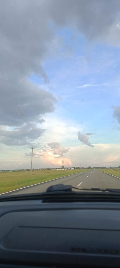 This media file captures a scenic view from inside a vehicle, looking out at a straight asphalt road stretching into the distance under a vast sky. The scene suggests a journey or travel. The road is marked with white lines and bordered by a green field on the left. Several utility poles are visible along the side of the road. In the field, a line of white structures, possibly tents or temporary shelters, can be seen in the distance, along with a few scattered trees and houses.
The sky is a prominent feature, filled with a dramatic interplay of clouds. Large, heavy gray clouds dominate the upper left portion of the frame, hinting at possibly stormy weather or recent rain. Towards the center and right, the sky transitions to a lighter, clearer blue with scattered, wispy white clouds, some illuminated by a soft, warm light that suggests either sunrise or sunset. This warm glow on the clouds indicates a time of day when the sun is low on the horizon.
The view is framed by the interior of a car, with a dark dashboard visible in the foreground, particularly at the bottom and lower edges of the image. This perspective places the viewer within the moving vehicle, observing the passing landscape. The overall mood is peaceful and contemplative, enhanced by the expansive sky and the sense of open space.
No text is visible in the media file. The location is identified as Малышево, Russia.