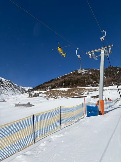 The image displays a sunny winter day at a ski resort in Schwendau, Austria. A yellow helicopter is airborne in the upper left quadrant, silhouetted against a clear blue sky. A prominent gray metallic ski lift pylon stands on the right, supporting overhead cables from which several empty ski lift chairs are suspended, one with a yellow support arm. The ground is extensively covered in groomed snow, forming ski slopes. A blue and yellow striped mesh safety fence stretches horizontally across the lower foreground, anchored into the snow. Adjacent to the ski lift pylon and fence are orange and blue protective pads. In the middle-ground, multiple structures, partially obscured by snow, are visible on a gentle slope. Distant mountains, featuring snow cover and patches of evergreen trees, form the horizon. Several small, indistinct human figures, consistent with skiers or snowboarders, are present on a slope in the far right-middle ground.