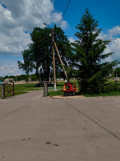 This is a medium shot, outdoor photograph taken on a sunny day in Velyka Bahachka, Ukraine. The scene is a paved road leading into what appears to be a park or recreational area. A large spruce tree dominates the right side of the frame, while a deciduous tree stands to the left behind a utility pole.

In the middle ground, a bright red, bench-like structure with a large golden bell sits next to the base of the spruce tree. Behind the bench, a tractor with a trailer is parked on a grassy area. Further in the background, there are bleachers or stadium seating, suggesting a sports or event venue. A decorative stone gate with an abstract design is visible on the left side of the road, leading into the grassy area.

The sky is blue with scattered white clouds. The overall mood is peaceful and rural. There is no visible text in the image. The time of day appears to be midday, given the bright sunlight and shadows.