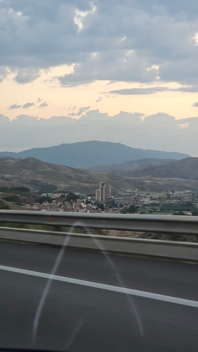 The image is a landscape view from a highway, looking towards a town nestled in a valley between mountains. The sky is partly cloudy, with areas of both blue sky and warm, golden-tinged clouds, suggesting it might be late afternoon or early evening. The sun appears to be setting or rising, casting a soft light over the scene.
The foreground of the image is dominated by the highway barrier and the asphalt road, with white lane markings visible. Beyond the barrier, the town of Morata de Jalón is visible, characterized by a cluster of buildings with red-tiled roofs. A prominent multi-story structure, possibly an industrial building or a tall residential complex, stands out within the town. The surrounding hills are dry and sparsely vegetated, typical of a Mediterranean climate. The mountains in the background are layered, with a hazy blue appearance due to distance.
There are no people visible in the image. The overall mood is peaceful and scenic, capturing a moment of travel through a picturesque rural area. The location cues strongly suggest Morata de Jalón, Spain, based on the topography and architectural style of the town. No visible text is present in the image.