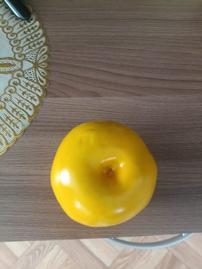 A single, bright yellow, possibly plastic, apple-shaped object sits on a dark wood grain surface. To the left of the object is a decorative, white and gold lace-like mat. The background is a wood grain surface with horizontal lines. A thin, light-colored curved object, possibly a chair leg, is visible at the bottom of the image against a parquet floor. The location of Шипуново, Russia, is not evident from the image.