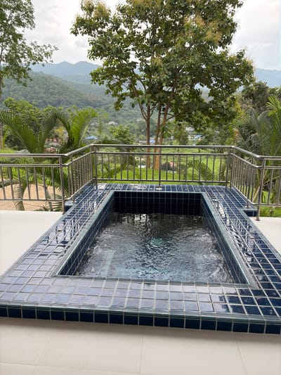 The image shows an outdoor hot tub on a tiled patio, with a metal railing overlooking a lush green valley with mountains in the background. The hot tub is rectangular and tiled with dark blue square tiles, and the water is rippling. The patio is tiled with light-colored square tiles. In the background, there are trees, palm trees, and some buildings in the distance. The sky is overcast. This scene is located in the city of Ban Kao Dua, Thailand.