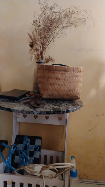 A still life arrangement is captured on a marbled-top, heart-cutout shelf unit. Dominating the scene is a woven bamboo basket filled with dried floral arrangements. On the marble surface, a dark rectangular object, possibly a notebook or tablet, sits next to a pair of scissors and a coiled reddish-brown rope. Below the shelf, in a white holder, are tangled blue wires, a checkered board, and white coiled cables, one with visible text. A partially visible plastic bottle with a blue cap sits to the right. The background is a textured, pale yellow wall. The scene is likely indoors, within a residence in Banjarsari, Indonesia, given the cultural context of woven bamboo baskets and the general domestic setting.