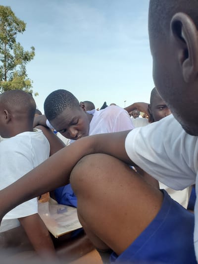 The image captures a group of young men outdoors, seemingly in a communal gathering or waiting area, possibly at Barakin Rafin Gora, Nigeria. The sky above is a pale blue, with a large tree visible on the left. The subjects are primarily focused on their immediate surroundings or on each other, with several looking down or away from the camera. They are dressed in white tops, suggesting a uniform or common attire. One individual in the foreground is leaning forward, with their head bowed, appearing tired or contemplative. Another person's arm is resting on their leg, showing a close proximity between individuals. In the background, other figures can be seen, some with their heads turned, indicating a shared space and potential interaction. The overall atmosphere appears relaxed and informal, with the natural light suggesting it's daytime.