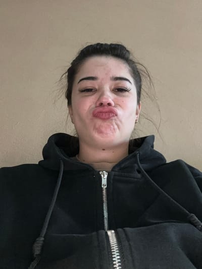 A young woman with dark hair pulled back in a bun and wearing a black zip-up hoodie is looking up at the camera with her lips puckered. She has long eyelashes and is wearing small hoop earrings. The background is a plain, textured wall. The image appears to be a selfie taken indoors. There is no indication of the city of Gavrilov Posad, Russia, in the image.