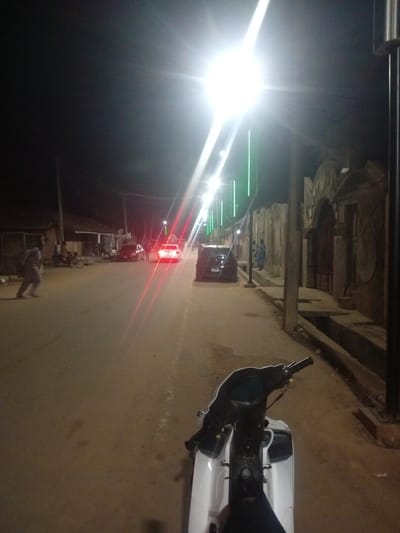 This media file depicts a nighttime scene on a street in Tundun Wada West, Nigeria, illuminated by bright streetlights and the taillights of vehicles. The foreground features the handlebars and front of a white scooter, suggesting the image was taken from the perspective of someone sitting on it or preparing to ride.

The street is paved and appears to be a main thoroughfare, with buildings lining one side. On the left, there are low-rise structures that appear to be shops or homes. On the right, a distinctive textured wall with an arched entrance gate is visible, hinting at a residential or commercial property.

Several people are present in the scene. A person in a light-colored garment is walking on the left side of the street. Further down the road, a person is standing near a motorcycle, and other individuals are seen near the buildings. Two cars are also visible: a dark sedan and a white sedan with its red taillights illuminated, indicating it is either parked or moving. Several green lights on poles are also visible, possibly decorative or functional lighting.

The time of day is clearly night, indicated by the darkness and artificial lighting. The weather appears clear. The scene conveys a sense of everyday life continuing into the evening in a Nigerian urban or semi-urban setting. No visible text is present in the image.