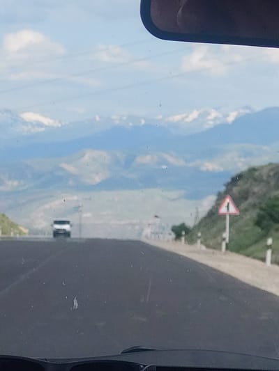 The image, oriented correctly, depicts a scenic view from inside a vehicle looking out over a mountainous landscape. On the far left, a portion of the vehicle's side mirror is visible. The right side of the frame shows a dark interior part of the vehicle, possibly a dashboard or window frame. The car window itself is speckled with small dust particles or water spots.
A bright blue sky with scattered white clouds dominates the upper half of the image. Below, a vast valley unfolds, flanked by multiple layers of green mountains and hills stretching into the distance. A dirt or gravel road runs along the right side of the frame, winding through the natural terrain.
In the mid-ground, a red-bordered white triangular warning sign with a black horizontal bar is prominently displayed on a white pole, positioned on what appears to be an embankment overlooking the valley. Further into the distance, within the valley, faint outlines of structures, possibly a remote settlement, can be seen. A small, light-colored vehicle, likely a car or van, is also visible on the road in the distant part of the valley.
The scene captures a daytime journey through a rural, mountainous region, consistent with the location context of Yelpin, Armenia. The weather appears clear and sunny. No explicit text is visible on the sign or elsewhere.
