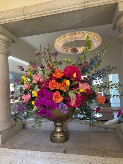 An ornate, multi-colored floral arrangement is presented in a large, polished brass urn-shaped vase, situated centrally on a textured light gray stone ledge. The arrangement features a dense mix of flowers including magenta phalaenopsis orchids, orange roses, pink roses, yellow tulips, hot pink hydrangeas, purple alliums, blue delphiniums, and various white and green filler foliage. The stone ledge is part of a larger architectural structure, flanked by a rounded, fluted stone pillar on the left and capped by a stone lintel overhead. In the background, a large, circular, crystal-encrusted chandelier hangs illuminated from a white ceiling. Beyond the floral display and stone architecture, an expansive, well-lit interior space is visible, containing a light-colored upholstered chaise lounge or sofa with visible stud detailing, and a distant light-colored door or window unit. No people, actions, or direct interactions are visible. The scene is located in McAllen, United States of America.