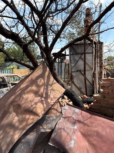 The image depicts a scene of extensive destruction in Nedryhailiv, Ukraine, under a clear blue sky with scattered white clouds. In the foreground, multiple large, warped, rust-colored metal sheets, exhibiting scorch marks and dents, are piled upon each other. Shadows from tree branches are cast across these sheets. To the left and slightly behind these sheets, the burnt-out, skeletal frame of a vehicle, consistent with a passenger car, is visible. It is blackened and stripped of its interior and exterior components, with only the shell remaining.

Dominating the mid-ground is a large, deciduous tree with thick, dark, and mostly bare branches, many of which appear charred. One large, blackened branch or trunk extends diagonally downwards towards the lower right, resting on a pile of fragmented red bricks and rubble. To the right of the tree, the remains of a brick structure are visible, consisting of a partially collapsed red brick wall and an exposed doorway frame with a grey metal gate. This gate features a diagonal cross-brace design. Beyond the immediate debris, a more intact, light-colored wooden fence is visible in the distant background to the left. No individuals or animals are present in the scene. The overall environment indicates the aftermath of significant damage or a destructive event.