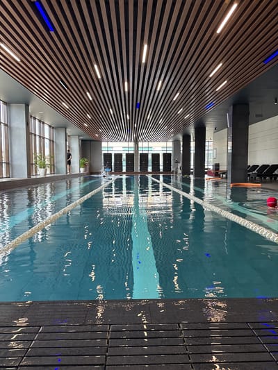 The image shows an indoor swimming pool in a modern facility, possibly located in the city of Monolit, Russia. The pool has clearly marked lanes separated by ropes. The water is a vibrant blue and reflects the overhead lighting and the architectural elements of the ceiling. The ceiling itself is a prominent feature, constructed with parallel wooden slats, interspersed with linear light fixtures and some blue accent lights. Large windows line one side of the pool area, allowing natural light to enter and providing a view of an urban environment.

On the left side of the pool, near the windows, are two potted plants. A person, dressed in dark shorts and a t-shirt, stands facing the pool and looking towards the far end. Further down the pool, several black rectangular fitness equipment units are visible, arranged in a row. Along the opposite side of the pool, beyond the lanes, a row of dark-colored lounge chairs is set up. In the foreground, the pool deck is made of dark, textured tiles. A red ball floats on the surface of the water towards the right side of the image. The overall impression is one of a clean, well-maintained, and contemporary fitness or leisure facility.