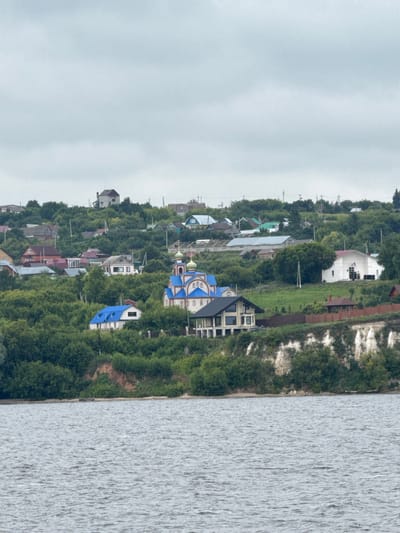 This is a medium-shot, landscape photograph taken from a body of water, likely a river, looking towards a densely vegetated hillside dotted with houses and a prominent church. The sky is overcast and a muted gray. In the foreground, the water exhibits small ripples. The hillside rises steeply, covered in a thick canopy of green trees and shrubs. Scattered amongst the foliage are various residential structures of different sizes and architectural styles, many with colorful roofs. A white building with a red roof is visible on the right side of the frame, and a distinctly blue-domed church with gold accents is centrally located on the hillside. A modern-looking house with a dark roof and large windows is situated closer to the bottom of the hill, near the water's edge. The hillside itself appears to have sections of exposed earth and light-colored rock formations. The overall impression is of a rural or semi-rural settlement nestled into a natural landscape. The location is identified as Krasnovidovo, Russia.