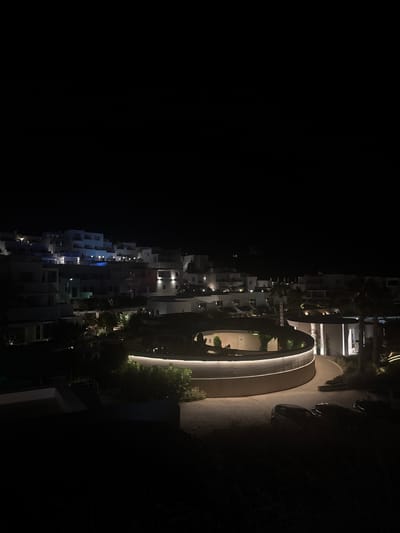 The image depicts a nighttime view of a modern resort complex situated on a hillside in Naousa, Greece. The architecture is characterized by white, cubist-style buildings with clean lines and minimal ornamentation, typical of Cycladic island design. Strategically placed outdoor lighting illuminates the structures and pathways, creating a warm and inviting ambiance.

In the foreground, a circular, multi-level building with a prominent curved facade is illuminated by soft, continuous lighting along its edges. This structure appears to be a central feature of the resort, possibly housing amenities or guest rooms. The surrounding landscape features manicured greenery and palm trees, also highlighted by subtle lighting.

Further up the hill, a cluster of white buildings cascades down the slope. These appear to be guest accommodations, with balconies and windows visible. The lighting here is more dispersed, with individual lights illuminating specific areas of the buildings and their entrances.

In the lower right portion of the image, several dark-colored cars are parked on a paved surface. The overall scene suggests a luxurious and contemporary resort designed to blend harmoniously with the natural topography and the iconic aesthetic of the Greek islands. The dark sky and artificial lighting emphasize the architectural forms and create a dramatic contrast.