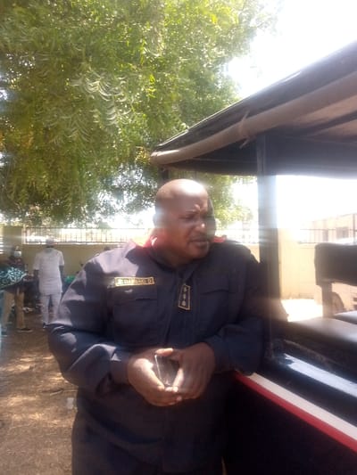 An outdoor scene in Kagoma, Nigeria, captured during daytime under bright sunlight. The primary subject is an adult male with a bald head and a mustache, positioned slightly right of center. He is wearing a dark blue uniform with a name patch on his left chest reading "DANSBAKI D" and a rank insignia consisting of three gold symbols vertically aligned on his right chest. A red trim is visible around his collar. He holds a black smartphone in both hands at waist level and is leaning against a dark-colored vehicle or structure that features a red and white stripe. His head is turned to the right, with his mouth slightly open, suggesting he is speaking or about to speak. In the background, to the left, are at least three other individuals; one is distinctly visible in a white traditional outfit and white cap, while another to his left appears to be wearing dark clothing and a face mask. The ground is bare earth. Above and behind the main subject, a dark roof structure with a gutter is visible, alongside extensive green foliage from a large tree. A fence or wall is discernible in the far background with faint structural outlines beyond it.