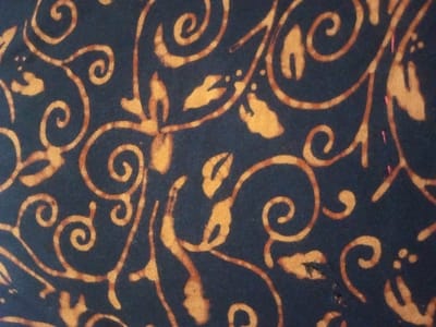 A close-up reveals a fabric with a swirling, botanical pattern. Tan-colored tendrils and leaves cascade across a dark blue background. The design appears to be a batik print, evidenced by the variations in tone and texture within the tan portions. The overall effect is intricate and somewhat organic, evoking a sense of nature. A few pink stitches are also visible on the fabric.