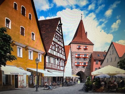 The image shows a street scene in Ellwangen, Germany, specifically the Schulkellerberg gate and surrounding buildings. The gate, a prominent stone structure with red-and-white window shutters, is centrally located. To its left, a half-timbered building with a steeply pitched, tile-covered roof features decorative wooden beams. Further left, a bright yellow building with multiple arched windows and a flat roof is visible.
In the foreground, several shops with awnings line the street. One awning displays the text "haarmoden Liedtke". A traditional street lamp stands in front of the shops. Benches are placed along the walkway, with a few people seated on them. Outdoor cafe seating with umbrellas is set up in front of other establishments, with patrons seated at tables. A bicycle is parked near the cafe area. The sky is blue with scattered white clouds. The overall impression is of a charming, historic European town square.