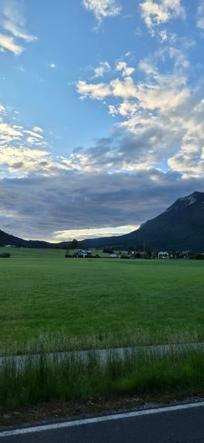 The image displays a rural landscape in Inzell, Germany, under a late afternoon or early evening sky. The upper third of the frame shows a sky with patches of light blue interspersed with numerous stratocumulus and cumulus clouds, some illuminated along their edges, suggesting indirect sunlight. To the right, a large, dark-green forested mountain with a distinct light-colored rocky peak dominates the horizon. To the left, a range of gentler, tree-covered hills forms a lower horizon line. The middle ground and foreground consist of extensive, vibrant green fields or pastures. Scattered across these fields and at the base of the hills, a small collection of detached houses and farm buildings, characterized by pitched roofs and varying colors, form a dispersed settlement. The immediate foreground reveals a dark asphalt road with a continuous white line marking its edge, bordered by a strip of tall green grass. No individuals or discernible activities are present in the scene.