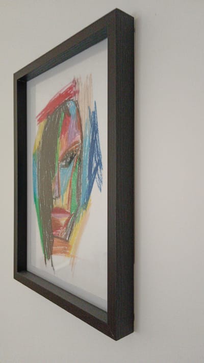 A framed, abstract pastel drawing of a face is mounted on a light-colored wall. The drawing features bold, fragmented lines in various colors, including red, blue, green, yellow, and brown, to depict facial features such as eyes, a nose, and lips. The frame is dark brown and appears to be made of wood. The perspective is from the side, showing the thickness of the frame and its placement against the wall. The lighting is soft and diffused, casting no strong shadows. The background is a plain, untextured wall, offering no specific environmental context. There is no indication of location in Sankt Pölten, Austria.