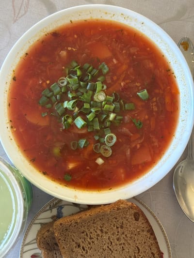 An overhead shot shows a bowl of borscht garnished with chopped green onions, along with two slices of dark bread on a decorative plate. A spoon rests to the right of the bowl, and a small green dish is visible to the left. The setting appears to be a table covered with a light-colored tablecloth. The image does not provide any information about a specific location in Rezekne, Latvia.