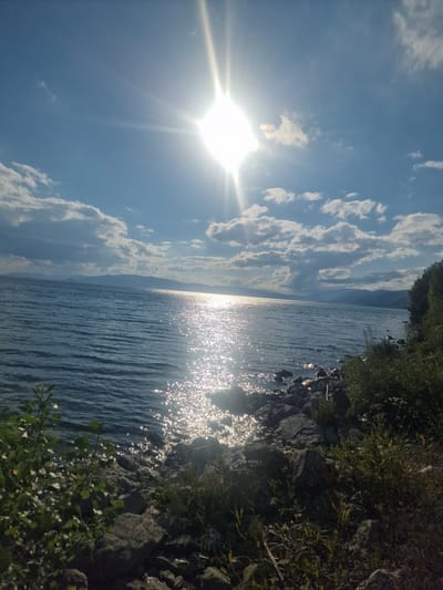 The image depicts a serene lake scene under a bright, sunny sky, likely taken in Sevan, Armenia. The sun is a dominant element, positioned on the left side of the frame and shining brightly with visible rays, suggesting it's midday or late afternoon. Scattered, fluffy white clouds dot the clear blue sky, adding texture and depth. The sun's reflection sparkles intensely on the calm surface of the water, creating a dazzling pathway of light that stretches towards the horizon.
The lake occupies the majority of the right side of the image. Its water appears dark blue and rippled, reflecting the light and the surrounding landscape. Along the edge of the lake, on the right, is a rocky shore with sparse green vegetation growing amongst the stones. The rocks are varied in size and texture, and some appear wet from the lapping water. In the distance, faint outlines of mountains are visible against the horizon, partially obscured by the haze or distance, adding a sense of scale and depth to the landscape. The overall mood of the image is peaceful and tranquil, evoking a sense of natural beauty and calm. There is no visible text in the image.