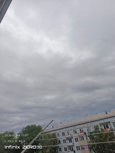 The image shows a cloudy sky over a city. In the foreground, there is a clothesline with a rusty pole extending upwards. Behind the clothesline, there is a light blue apartment building with multiple windows and balconies. Trees with green foliage are visible between the clothesline and the building. Text overlays indicate the date "2026/07/14 17:07", coordinates "E 131,3 N 44,3", and "Infinix ZERO30". Based on the coordinates provided, the scene is located in Letno-Hvalinskoe, Russia. There are no people visible in the image.