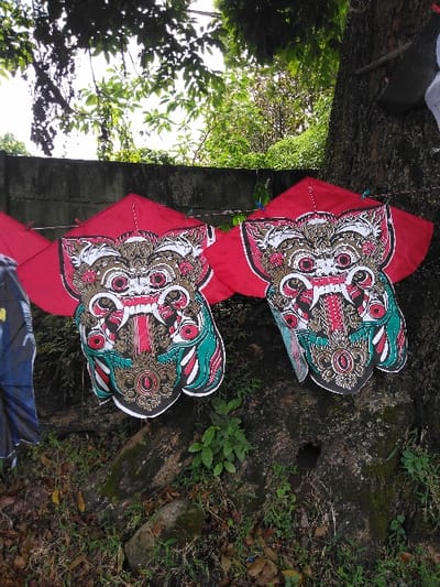 The scene depicts an outdoor display of several kites in Thit Seint Pin, Myanmar. Two primary kites are prominently featured in the center-right, suspended from lines tied to a large tree trunk on the right and extending towards a weathered grey concrete wall in the background. These two kites are identical, each featuring a red, roughly diamond-shaped upper section and a lower section designed as a stylized, intricate face with elements in white, black, red, green, and gold, characterized by large eyes and visible fangs. A partial red kite is visible to the immediate left of these, and a partial blue kite is further to the left. The ground beneath the display consists of visible tree roots, sparse green vegetation, moss, and scattered brown leaf litter. Dense green foliage and tree branches are visible above the wall and tree trunk, obscuring the sky. No individuals are present in the image.