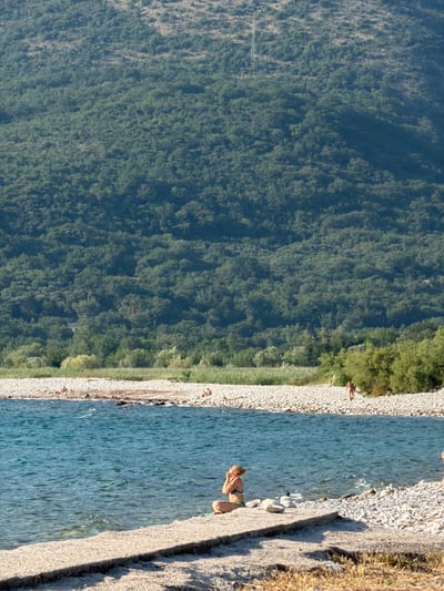 This is a vertical, eye-level shot of a rocky beach next to a body of water, with a densely forested mountainside in the background. The scene appears to be set in Buljarica, Montenegro, under bright daylight. The water is a clear, tranquil blue, with gentle ripples on its surface. The beach is composed of smooth, pale grey pebbles.

Several people are visible along the shoreline. In the foreground, a woman in a bikini is seated on the rocky bank, looking downwards and holding her hands near her face. Further along the beach, other individuals are scattered, some appearing to be standing or walking. One person is seen in the water near the shore, and another is sitting further up the beach.

The mountainside behind the beach is covered in lush, dark green foliage, suggesting a dense forest. The lighting indicates it is daytime, with sunlight casting shadows and illuminating the scene. The overall atmosphere is relaxed and summery, with people enjoying the natural environment by the water. There is no visible text in the image.