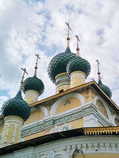 The image shows the domes and upper facade of an Orthodox church against a cloudy sky. The church has multiple green, onion-shaped domes, each topped with a cross. The facade is painted yellow and features decorative trim and architectural details in white and green. The overall scene is a religious architectural landmark. The location is identified as Vasil'ki, Russia.