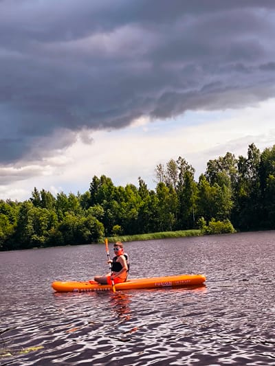 A person is paddleboarding on a lake in Vecumnieki, Latvia. The person is wearing a life vest and sunglasses. They are holding a paddle and are in the middle of the lake. The lake is surrounded by a forest of green trees. The sky is cloudy and appears to be overcast. The water is calm with ripples. The paddleboard is orange and has text on it that says "AQUA MARINA FUSION 10'10 x 32".