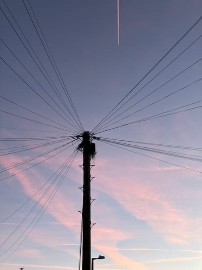 The image shows a telephone pole with numerous wires extending from it in various directions, silhouetted against a twilight sky. The sky transitions from a pale blue at the top to pink and purple hues lower down, with wispy clouds. An airplane's contrail is visible high in the sky. The telephone pole is dark and appears to be made of wood, with electrical boxes and other equipment attached near its top. In the lower part of the image, a streetlamp is visible on the right, and what appears to be a small aerial or antenna structure is on the left, closer to the bottom edge. The scene depicts a common urban or suburban utility infrastructure against a backdrop of a colorful sky, suggesting either dawn or dusk. The location is Aveley, United Kingdom.