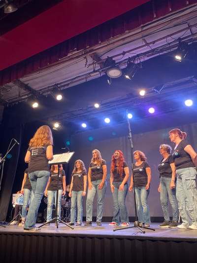 A group of women, likely a choir, are performing on a stage under theatrical lighting. The individuals are dressed in matching black t-shirts with a circular logo that appears to read "Gospel" with a heart symbol, and blue denim jeans. One woman, positioned towards the left of the frame and facing away from the camera, seems to be leading the group, holding a music stand. The stage is set up for a performance, with microphones, lighting equipment, and a drum set visible in the background. The backdrop appears to be a dark curtain or screen. The overall atmosphere suggests a live musical event, possibly a gospel concert, taking place in a performance venue. The image was taken in Arenys de Mar, Spain.
