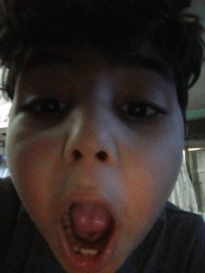 The image is a close-up selfie of a young boy with dark hair. He is wearing a dark shirt. His mouth is open wide, and he is looking directly at the camera. The boy's face is illuminated by an artificial light source, likely a cell phone, casting an uneven light across his face. The image was taken in a residential area in Alexandria, Egypt, at coordinates 31.227 latitude and 29.962 longitude. The background is out of focus, but it appears to be a room with a curtain in the background. There is no other identifiable detail about the environment.