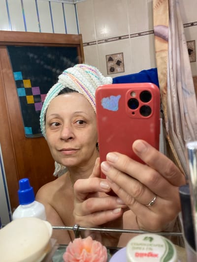 A woman, wearing a towel turban and no visible clothing on her upper body, takes a selfie in a bathroom mirror. She holds a red smartphone in her right hand, capturing her reflection. Various bathroom items are visible in the foreground and background, including bottles of toiletries, a bar of soap, and what appears to be a decorative soap flower. The bathroom walls are tiled, and a shower curtain is partially visible. The scene does not provide any specific details to indicate a location in Yokohama, Japan.