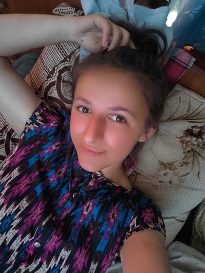 The media file is a close-up, overhead shot of a young woman lying down, looking at the camera with a slight smile. She has her hair pulled up in a bun and is resting her head on her arm. She is wearing a colorful, patterned shirt with blue, pink, and black designs. The background appears to be a bed or couch with patterned fabric. The lighting suggests it is daytime, possibly indoors. The woman's expression is relaxed and amiable. There is no visible text in the image.
