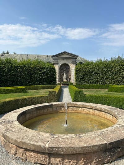 The image shows a formal garden with a circular stone fountain in the foreground. The fountain has a single jet of water spraying upwards from its center. The water in the fountain is a murky yellow-brown color. The fountain is surrounded by gravel. Behind the fountain, a gravel path leads through manicured hedges towards an arched entryway. Within the entryway, a pale stone statue of a human figure stands. Tall, dark green hedges form walls on either side of the garden, and a building with a dark, sloped roof is visible in the background. The sky is blue with scattered white clouds. This is the garden of the Náchod Castle in Náchod, Czechia. There are no people visible in the image.