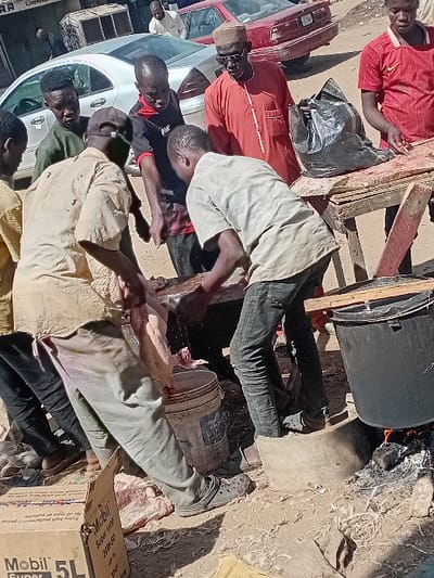 The image captures an outdoor scene in Sharada, Nigeria, depicting the processing of a freshly slaughtered animal, likely a goat or sheep, surrounded by approximately eight males. In the foreground, two men are actively engaged with the animal, which lies on a low surface or the ground; the man on the left wears a light-colored shirt and dark pants, while the man on the right wears a light gray shirt and dark jeans, both bending over the carcass. Another man in a black and red shirt also has his hands on the animal in the center.
Several other individuals observe the activity: a man in a green shirt to the left, a man in a red Nike-branded shirt standing behind a makeshift wooden table to the right, and a man in a red traditional outfit with a brown cap and sunglasses in the mid-ground. A large black plastic bag rests on the wooden table. To the far right, a large black cooking pot is positioned over an open fire, supported by concrete blocks. Two plastic buckets, one white and one brown/red, are visible on the ground near the working men. The ground surface is unpaved dirt, visibly scattered with animal hair and debris. A "Mobil Super 5L" cardboard box is situated in the lower-left corner. In the background, a silver Mercedes-Benz sedan and a maroon car are parked on what appears to be a street adjacent to commercial buildings.