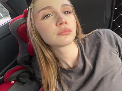 The image shows a young woman with blonde hair and makeup, looking upwards and to her right. She is in what appears to be the interior of a vehicle, possibly a car, as indicated by the presence of a car seat with red and black fabric, and a dark padded headrest. Her head is resting on something, and her expression is relaxed or perhaps tired. The background suggests an outdoor environment with trees and a road visible through a window, likely from the perspective of someone seated in the back of the car. There is no direct information indicating the specific location of Новая Отрадовка, Russia, within this image.