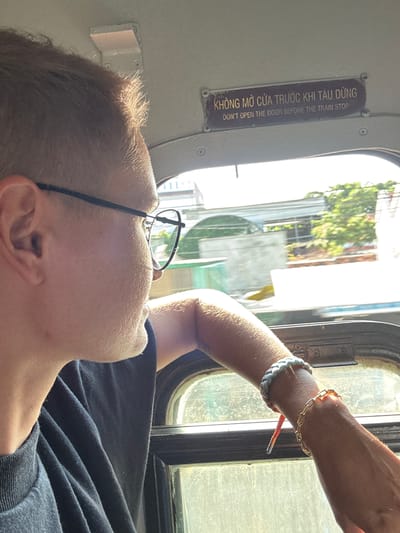 The image shows a close-up of a person with short, light brown hair and glasses, looking out of a train window. The person is wearing a dark t-shirt. Their right arm is resting on the window frame, and they are wearing two bracelets on their wrist. Outside the window, there are buildings and trees, suggesting an urban or semi-urban environment. A sign above the window reads, "KHÔNG MỞ CỬA TRƯỚC KHI TÀU DỪNG DON'T OPEN THE DOOR BEFORE THE TRAIN STOP". This sign indicates that the image was taken on a train in Vietnam. The location is specified as Vạn Giã, Vietnam.