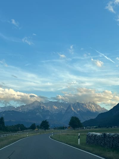 The image shows a winding asphalt road leading towards a majestic mountain range. The sky above is a clear blue with scattered white clouds. The mountains, with their rugged peaks, are partially obscured by soft, white clouds, giving them a dramatic appearance. The lower slopes of the mountains are covered in dense green forests. In the foreground, the road is flanked by grassy fields. To the right of the road, a neatly stacked pile of firewood, covered with a dark tarp, is visible. Several white posts with black markings are placed along the side of the road. In the middle ground, there are scattered trees and some signs of human infrastructure, including what appears to be a guardrail and some road signs. The overall scene depicts a serene and picturesque landscape, likely in the vicinity of Farchant, Germany, known for its proximity to the Bavarian Alps.