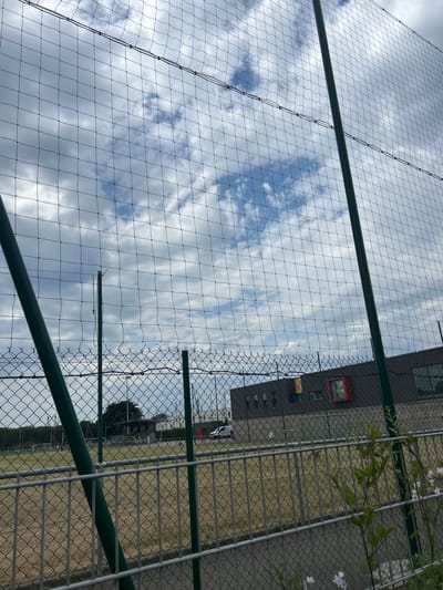 The image shows an outdoor scene on a cloudy day, viewed through a protective net fence. In the foreground, a green metal pole is visible on the left, and a grey metal fence runs horizontally across the lower portion of the image. Beyond the fences, a sports field with dry grass is visible, with goalposts and lights indicating it is likely a football (soccer) pitch. In the midground, a modern building with dark walls and a distinctive red window or architectural feature is situated behind the field. A white van is parked near the building. In the background, trees and other structures are visible under a sky filled with grey and white clouds. The overall impression is of a sports facility or school complex in an urban or suburban setting in Saint-Nazaire, France. There are no people clearly visible in the image.