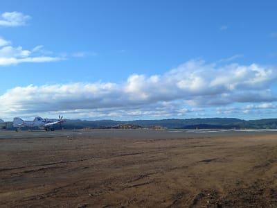 The scene depicts an airfield in Santa Juana, Chile, under a clear blue sky with scattered cumulus clouds. In the midground, a white single-engine turboprop aircraft, bearing the registration "CC-DOJ" and featuring red accents, is stationary on a paved tarmac or runway, oriented towards the left. A portion of a white building is visible behind the aircraft's left wing. The foreground consists of a wide area of reddish-brown, disturbed earth, indicative of dry soil or dirt. To the right of the aircraft, the ground appears as a lighter-colored, dry, barren expanse. The background is composed of distant, dark green forested hills, with visible piles of timber or wood debris situated between the midground and the distant hills. No persons are present, and no activity is observed.