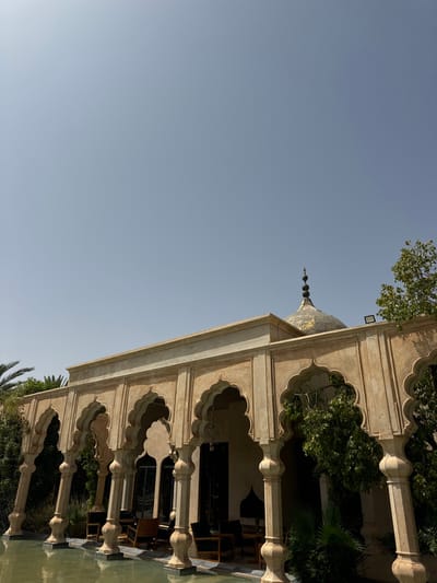 The image shows a courtyard, framed by a series of arches, located at the coordinates 31.664 latitude and -7.871 longitude in Ouled Jelal, Morocco. The courtyard is situated within a building that features a dome with a pointed spire. Lush foliage surrounds the courtyard, and the arches create a shaded, tranquil space. The arches are intricately decorated with traditional Moroccan patterns, and the courtyard floor is covered with a shallow pool of water. There is a smattering of chairs in the courtyard, suggesting a place for relaxation and contemplation. The sky above the courtyard is a clear, pale blue, with no clouds visible. The scene is serene and peaceful, reflecting the quiet beauty of traditional Moroccan architecture.