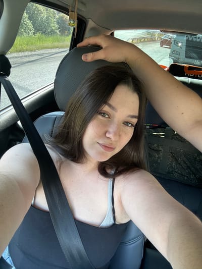 A woman with dark hair and green eyes is taking a selfie in the passenger seat of a car. She is wearing a black tank top and a grey bra. A seatbelt is across her chest. A hand is visible resting on the back of her headrest, suggesting another person is in the car. Through the car window, a road and trees are visible. In the background, a truck is driving on the road. The scene appears to be in Pesterevo, Russia.