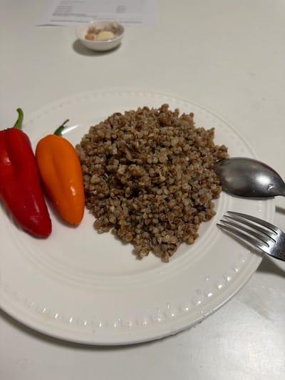 A white plate holds a serving of cooked buckwheat and two small, colorful bell peppers, one red and one orange. A fork and spoon are positioned to the right of the plate. In the background, a small white bowl contains what appears to be butter and garlic. A document is visible on a white surface behind the bowl. The scene is set indoors, on a white table. This image was captured in Merrifield, United States of America.