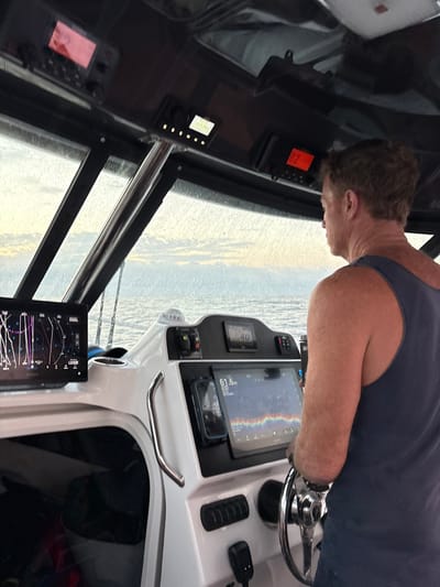 A person steers a boat, visible from the shoulders up. The captain grips a chrome steering wheel. The dashboard features multiple screens displaying navigational information and speed. One screen shows sonar data. Rain streaks the window, partially obscuring the view of the water. The boat's interior is modern, with a white and black color scheme.